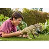 Na zdjęciu znajduje się figurka dinozaura na trawie, otoczona roślinami. W tle widoczny jest żywopłot oraz fragmenty nieba.