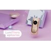 Na zdjęciu znajduje się depilator BEAUTIFLY LumiThera Epil IPL w kolorze różowym, obok którego widnieje napis "LumiThera epilseries". W tle widoczne są elementy dekoracyjne w odcieniach fioletu i różu oraz pudełko z roślinnym wzorem.