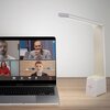 Na zdjęciu znajduje się otwarty laptop z widocznym ekranem, na którym trwa wideokonferencja z czterema osobami. Obok laptopa stoi biała lampka biurkowa o nowoczesnym designie, z widocznym symbolem plusa na podstawie.