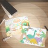 Na zdjęciu znajduje się lampka biurkowa oraz dwa kolorowe rysunki przedstawiające domek z ciastkiem i dinozaury w otoczeniu palm i tęczy. Obok rysunków leżą dwa markery, jeden z widocznym napisem "OREX".