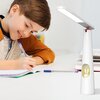 Na zdjęciu znajduje się biała lampka biurkowa o nowoczesnym designie, z wbudowanym elementem dekoracyjnym w kształcie króliczka. Obok lampki widoczna jest osoba pisząca w zeszycie.