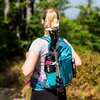 Na zdjęciu widoczny jest turkusowy plecak z napisem "NILS CAMP" oraz "NC1950", do którego przymocowane są kijki trekkingowe i czarny bidon z napisem "NILS". W tle znajduje się rozmyte, zielone otoczenie, sugerujące leśne środowisko.