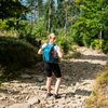 Osoba stoi na kamienistej ścieżce w lesie, nosząc turkusowy plecak z widocznym napisem "NILS CAMP". W tle widać drzewa i zieloną roślinność.