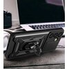 Na zdjęciu znajduje się czarne etui na telefon z widocznym uchwytem i trzema otworami na aparaty, leżące na drewnianym stole. W tle widać częściowo otwarty laptop oraz słuchawki.