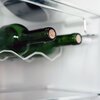 Na zdjęciu widać wnętrze lodówki z metalowym stojakiem na butelki, na którym leżą dwie zielone butelki z korkami. Tło jest jasne, a elementy lodówki mają metaliczne wykończenie.