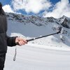 Osoba trzyma czarny uchwyt selfie z zamocowaną kamerą na tle ośnieżonych gór. Uchwyt ma pasek na nadgarstek i jest rozciągnięty, a w tle widać niebo z chmurami.