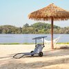 Na zdjęciu znajduje się szary leżak turystyczny z daszkiem, ustawiony na piaszczystej plaży. W tle widoczny jest parasol z trzciny oraz hamak, a za nimi rozciąga się spokojna woda i zalesiony brzeg.