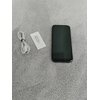 Na zdjęciu znajduje się smartfon z ciemnym ekranem, obok którego leży zwinięty biały kabel oraz metalowy kluczyk do tacki SIM na białym tle. Wszystkie przedmioty umieszczone są na szarej powierzchni.