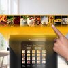 Na zdjęciu widoczny jest multicooker z wyświetlaczem dotykowym, na którym pokazane są różne programy gotowania z obrazkami potraw. Logo "TurboTronic Kitchen Electronics" znajduje się nad wyświetlaczem, a palec wskazuje na jeden z programów.