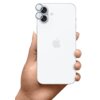 Na zdjęciu widoczny jest biały smartfon trzymany w dłoni, z logo w kształcie nadgryzionego jabłka na tylnej obudowie. W górnej części urządzenia znajdują się dwa obiektywy aparatu z nałożonymi srebrnymi nakładkami ochronnymi.