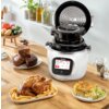 Na zdjęciu znajduje się multicooker marki Tefal z otwartą pokrywą, ustawiony na drewnianym blacie, obok talerzy z pieczonym kurczakiem i frytkami, makaronem z zapiekanką oraz kawałkami ciasta. Na ekranie urządzenia widoczny jest tekst: "What shall we cook