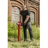 Mężczyzna w czarnym t-shircie i spodniach roboczych DEDRA BH54SP-M (rozmiar M/50) obsługuje czerwony hydrant na tle ceglanego budynku z dużymi oknami. Obok hydrantu znajduje się znak z literą "H".