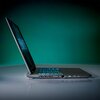 Na zdjęciu widoczny jest otwarty laptop z podświetlaną klawiaturą, ustawiony na gładkiej powierzchni. W tle znajduje się jednolite, zielone tło, a na ekranie laptopa widoczny jest nieczytelny tekst.