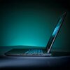 Laptop z otwartym ekranem, widoczny z boku, stoi na gładkiej powierzchni. Klawiatura jest podświetlona, a w tle znajduje się zielono-niebieskie oświetlenie.