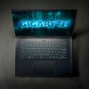 Na zdjęciu widoczny jest otwarty laptop z podświetlaną klawiaturą i dużym touchpadem. Na ekranie wyświetla się logo "GIGABYTE" w niebieskim kolorze na tle futurystycznej grafiki.