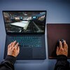 Laptop z podświetlaną klawiaturą wyświetla grę na ekranie. Obok laptopa znajduje się mysz na podkładce, a na klawiaturze i myszy spoczywają dłonie.