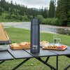 Niebieski termos NILS CAMP NC3803 o pojemności 500 ml stoi na składanym stole piknikowym, otoczony talerzami z przekąskami i szklanką wody. W tle widoczny jest zielony krajobraz z rzeką, drzewami i namiotem.