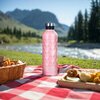 Różowa butelka termiczna o pojemności 750 ml stoi na czerwono-białym kocu piknikowym, na tle górskiego krajobrazu z rzeką i drzewami. Na butelce widnieje logo "NILS CAMP", a obok niej znajdują się koszyk z jedzeniem i drewniana tacka z przekąskami.