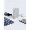 Na zdjęciu znajduje się szary powerbank z oznaczeniem "10000" na boku, otoczony przez różne urządzenia elektroniczne, takie jak smartfony, tablet i etui na słuchawki. Tło jest jasne, a produkty są rozmieszczone na płaskiej powierzchni.