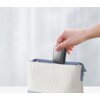 Na zdjęciu widać rękę wkładającą szary powerbank do jasnej torebki z niebieskimi elementami. Powerbank ma gładką powierzchnię i zaokrąglone krawędzie.