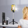 Czarny uchwyt selfie z zamocowanym smartfonem, na ekranie którego widoczna jest osoba na tle zielonego parku. Na szczycie uchwytu znajduje się okrągłe światło LED.