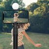 Na zdjęciu znajduje się uchwyt selfie w kolorze czarnym, trzymający smartfon, na którym widoczny jest obraz osoby w różowym płaszczu i kapeluszu, trzymającej kubek. W tle znajduje się jasnoniebieskie tło, a nad smartfonem umieszczone jest okrągłe światło.