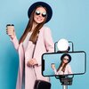 Czarny uchwyt selfie HURTEL WR1YXS jest zamontowany na rozkładanym statywie, a na nim znajduje się smartfon. W tle widać osobę siedzącą na kanapie, trzymającą filiżankę, a na stoliku leżą książki.