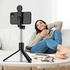 Na zdjęciu widoczny jest czarny uchwyt selfie z zamocowanym smartfonem, trzymany przez osobę w zimowym ubraniu. W tle znajdują się cztery osoby ubrane w zimowe kurtki i szaliki, pozujące do zdjęcia.