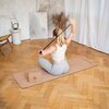 Na zdjęciu widać osobę siedzącą na macie do jogi w jasnym pomieszczeniu z drewnianą podłogą, trzymającą elastyczną taśmę nad głową. Na macie znajduje się prostokątny blok z widocznym logo i wzorem, a w tle stoją dekoracje i zasłony.
