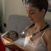 Osoba siedzi na łóżku, trzymając książkę, z lampką do czytania zawieszoną na szyi, emitującą światło na strony. Na lampce widoczny jest napis "Innovagoods".