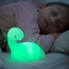 Na zdjęciu widać świecącą na zielono lampkę w kształcie dinozaura, umieszczoną na łóżku obok śpiącego dziecka. Lampka ma prosty, zaokrąglony kształt z czerwonymi i czarnymi detalami na głowie.
