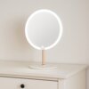 Na zdjęciu znajduje się okrągłe lusterko kosmetyczne z podświetleniem LED, umieszczone na blacie mebla. Lusterko ma prosty, nowoczesny design z drewnianą nóżką i białą podstawą.