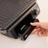 Czarny grill elektryczny z widocznym logo "BRAUN" na przedniej części, z pokrętłem regulacji temperatury i ręką otwierającą dolną szufladę. Pokrywa grilla ma żebrowaną powierzchnię.