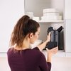 Na zdjęciu widać osobę wkładającą czarny grill elektryczny do szafki kuchennej, w której znajdują się białe talerze i miski. Produkt jest ustawiony pionowo, a jego powierzchnia jest gładka i ciemna.