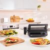 Na zdjęciu znajduje się czarny grill elektryczny z logo "Braun" na blacie kuchennym, obok którego leżą talerze z grillowanymi potrawami, w tym warzywami i mięsem. W tle widoczna jest jasna kuchnia z drewnianą deską do krojenia i miską z owocami.