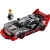 Na zdjęciu znajduje się model samochodu wyścigowego z klocków LEGO, z widocznymi napisami "Audi" i "e-tron" oraz figurka ludzika LEGO w kombinezonie, trzymająca kask. Samochód ma czarno-czerwone barwy z detalami w kolorze szarym i białym.
