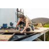 Na zdjęciu widać osobę pracującą na drewnianym tarasie, używającą klucza udarowego BOSCH Professional GDX 18V-210 C 06019J0202. W tle znajduje się dom, narzędzia oraz krajobraz z drzewami i wzgórzami.