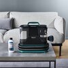 Na zdjęciu znajduje się odkurzacz Tefal Clean It z widocznym logo "Tefal Clean It" umieszczony na stoliku przed szarą sofą. Obok odkurzacza stoi butelka z płynem czyszczącym z etykietą zawierającą tekst i grafikę.