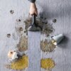 Ręka trzymająca odkurzacz czyszczący zabrudzony dywan, na którym znajdują się plamy i przewrócone naczynia z resztkami napojów. Odkurzacz tworzy czysty pas na dywanie, usuwając zabrudzenia.