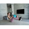 Na zdjęciu znajduje się osoba ćwicząca w nowoczesnym wnętrzu z szarymi ścianami, trzymająca kettlebell. W tle widoczna jest niebieska piłka do ćwiczeń, pomarańczowy wałek oraz urządzenie z napisem "HORIZON".