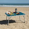 Na zdjęciu znajduje się niebieski stolik kempingowy z metalowymi nogami, ustawiony na piaszczystej plaży. Na stoliku leżą różne produkty spożywcze, w tym kanapki, owoce, winogrona, a także termos; widoczne jest logo "NILS" na krawędzi stolika.
