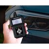 Osoba trzyma w ręku czarny tester diagnostyczny z wyświetlaczem, na którym widnieje tekst: "OBD2/EOBD Code Reader", a poniżej symbole i przyciski nawigacyjne. W tle widoczny jest fragment reflektora samochodowego.