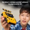 Na zdjęciu widoczny jest żółty model samochodu z klocków, trzymany w dłoni, z napisem "Na dzieci czekają wyścigi i zabawa w odgrywanie ról" na górze. W tle znajduje się osoba, której twarz jest nieostra.