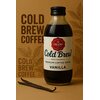 Butelka napoju "Cold Brew" o smaku waniliowym stoi na tle brązowego kartonu z napisem "COLD BREW COFFEE" i grafiką liści, obok leży laska wanilii. Na etykiecie butelki widnieje napis "GRANO TOSTADO", "100% Arabika", "Premium Coffee Drink", "Vanilla".