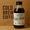 Butelka z napojem "100% Arabica Beans Coffee Cold Brew Brazylia" marki Grano Tostado o pojemności 200 ml stoi na tle kartonu z napisem "COLD BREW COFFEE" i grafiką liści. Etykieta butelki zawiera również logo Grano Tostado.