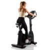 Na zdjęciu znajduje się rower elektromagnetyczny z napisem "HAMMER Maximum" na obudowie. Osoba siedzi na rowerze, trzymając kierownicę, ubrana w jasny top i ciemne spodnie sportowe.