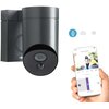 Na zdjęciu znajduje się antracytowa kamera zewnętrzna Wi-Fi marki Somfy, obok której widoczny jest smartfon z aplikacją pokazującą obraz z kamery. Na ekranie telefonu widać osobę stojącą na zewnątrz, a w tle znajdują się ikony przedstawiające człowieka, s