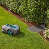 Na zdjęciu znajduje się robot koszący na trawniku, obok stacji dokującej, z widocznymi niebieskimi akcentami na obudowie. W tle widać żywopłot oraz fragmenty roślinności.