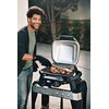 Na zdjęciu znajduje się otwarty grill elektryczny z kilkoma kawałkami mięsa na ruszcie, a obok stoi osoba trzymająca szczypce. Na grillu widoczne jest logo "Weber".