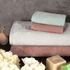 Na zdjęciu widoczne są trzy ręczniki w różnych kolorach ułożone jeden na drugim, obok znajdują się białe kwiaty, kostka mydła i flakonik perfum. Tło stanowi szara, teksturowana powierzchnia.